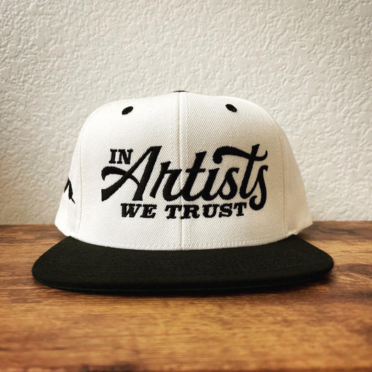 white and black hat on wood table with in artists we trust embroidery